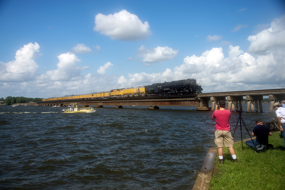 A steam locomotive hauling a yellow passenger train near a lake.