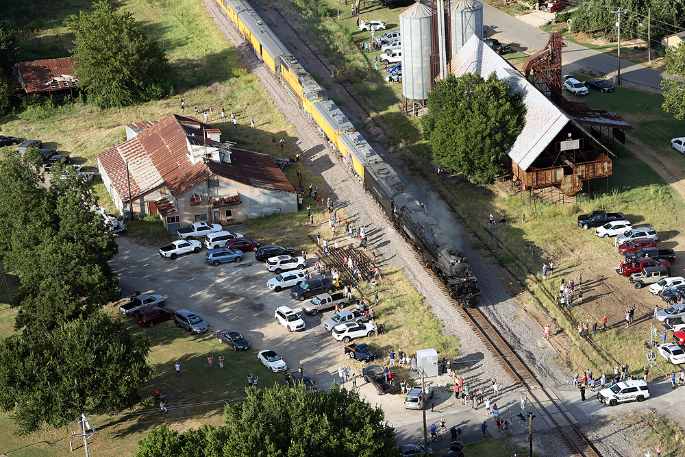 Steam locomotive passing between rural steel buildings a small crowd of people.