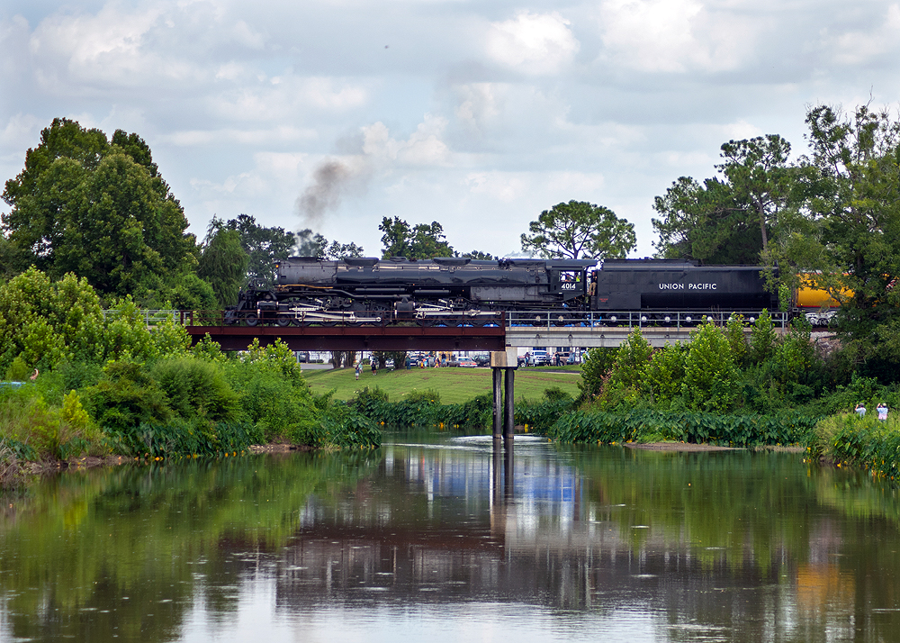 Large steam locomotive rolls over a short deck bridge framed by water, trees, and green shrubs.