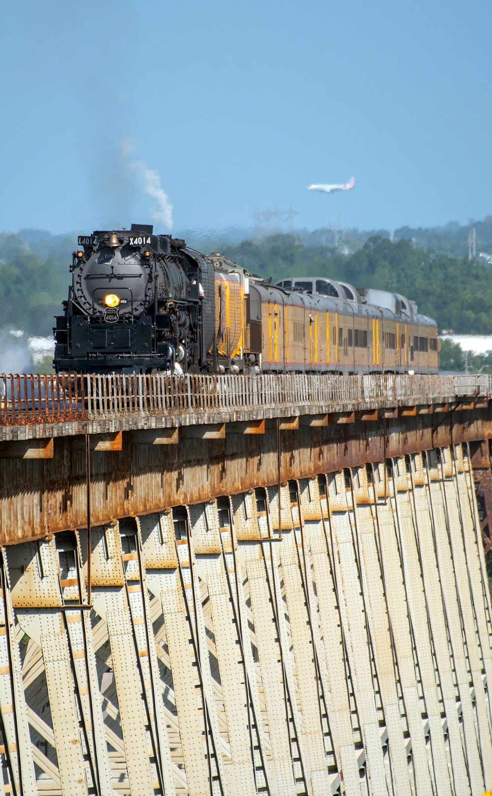 Large steam locomotive with train on a long bridge with a commercial jetliner in the distant background.