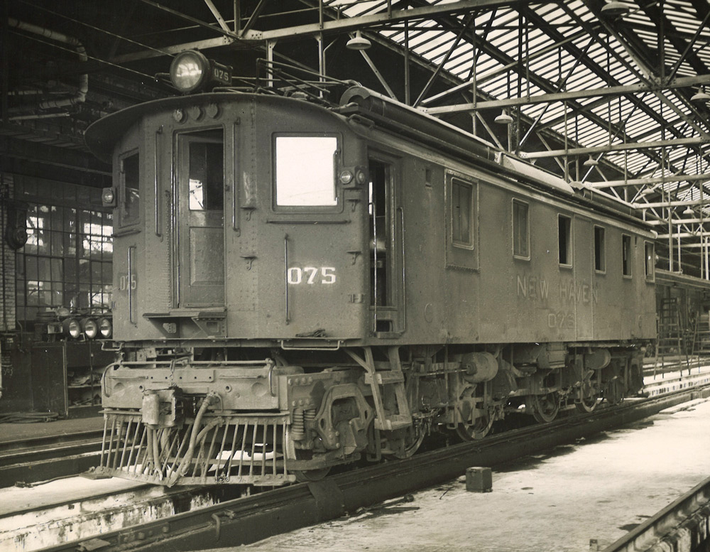 Black-and-white three-quarter-angle photo of box-cab electric locomotive inside shop building.