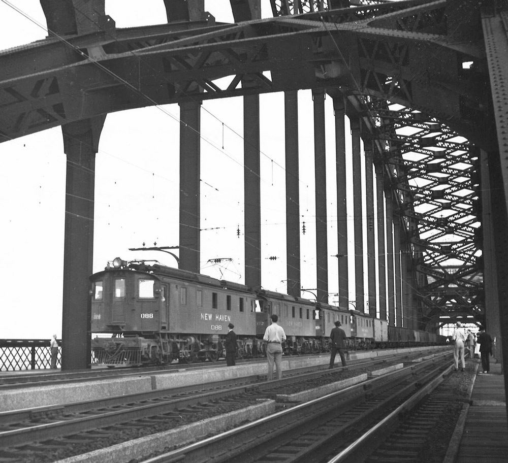 Black-and-white photo of people watching electric locomotives on bridge with freight train