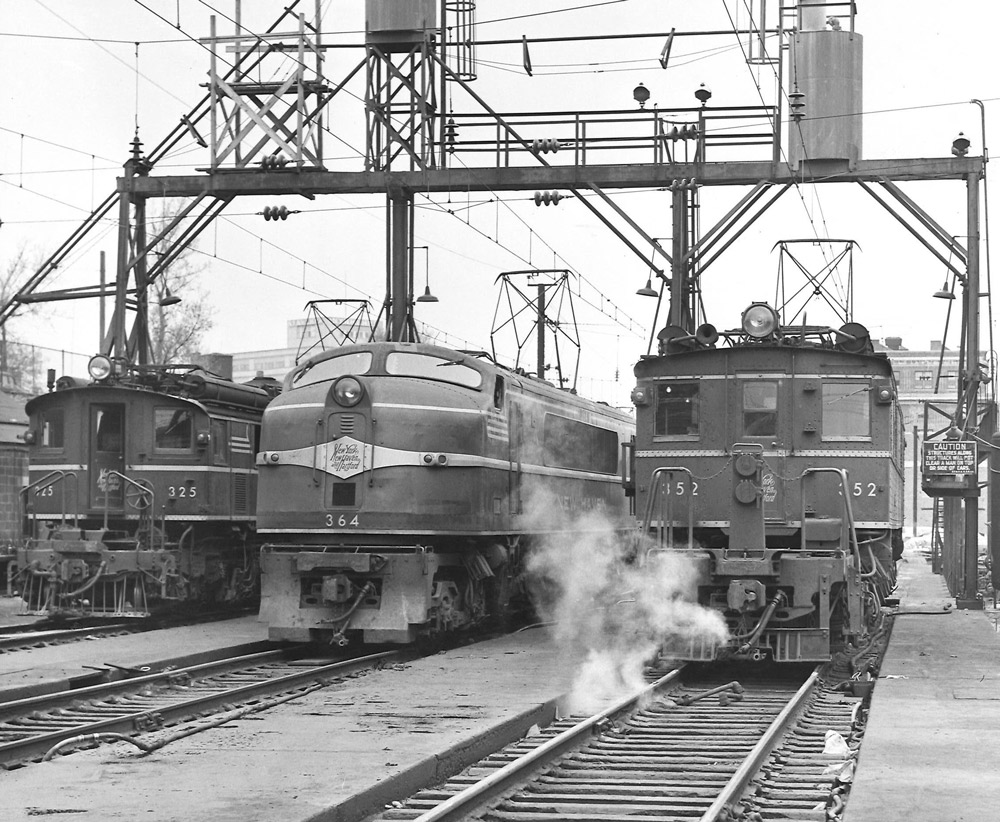 Black-and-white view of front ends of three electric locomotives on adjacent servicing tracks.