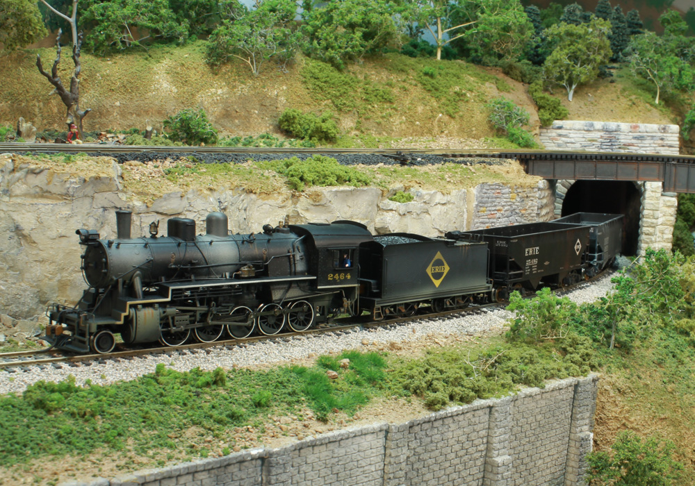 A steam locomotive leads a train out of a tunnel on the side of a wooded hill