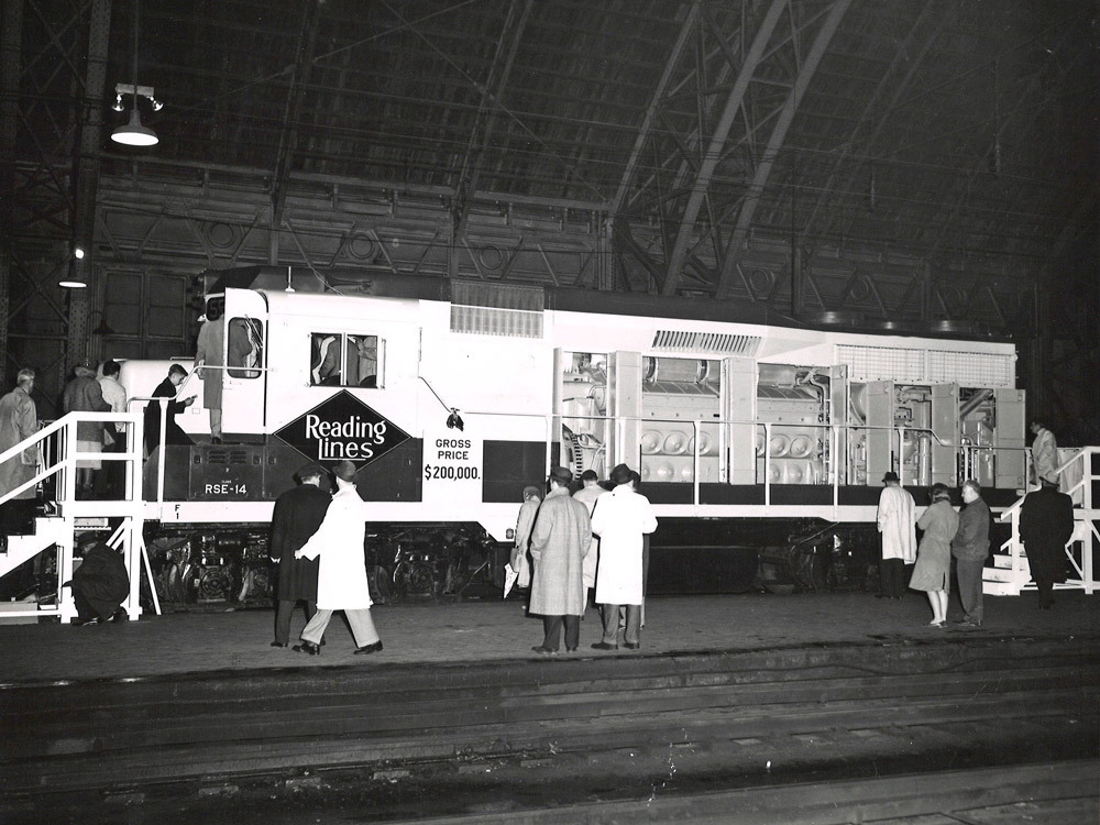 Black-and-white broadside photo of road-switcher diesel locomotive with hood doors open and people