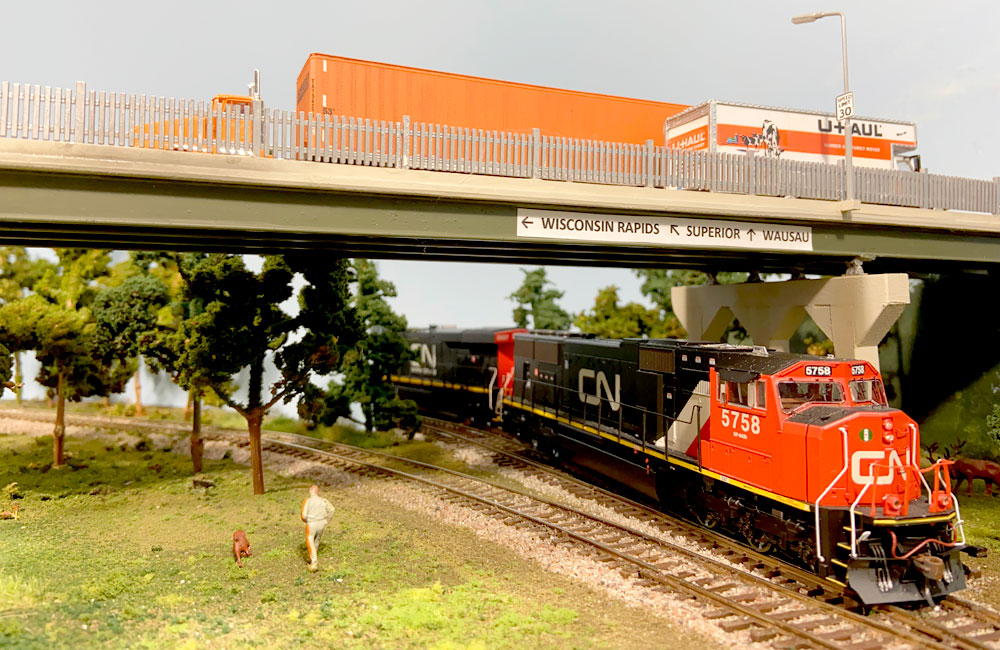 A pair of red-and-black diesels pass beneath a highway overpass