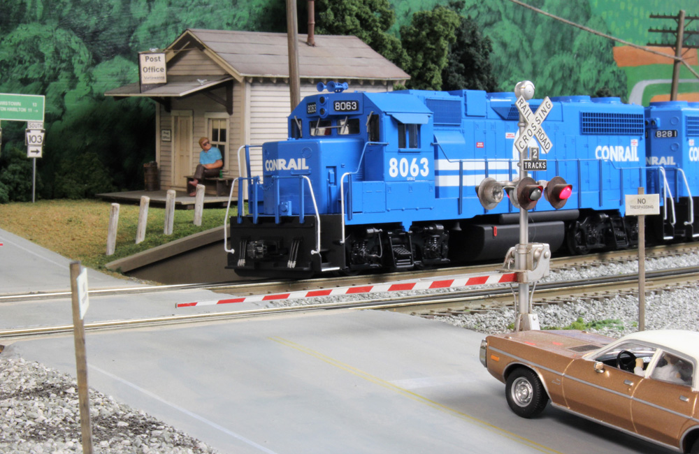 A bright blue Conrail diesel crosses a road in front of an old-fashioned white clapboard Post Office