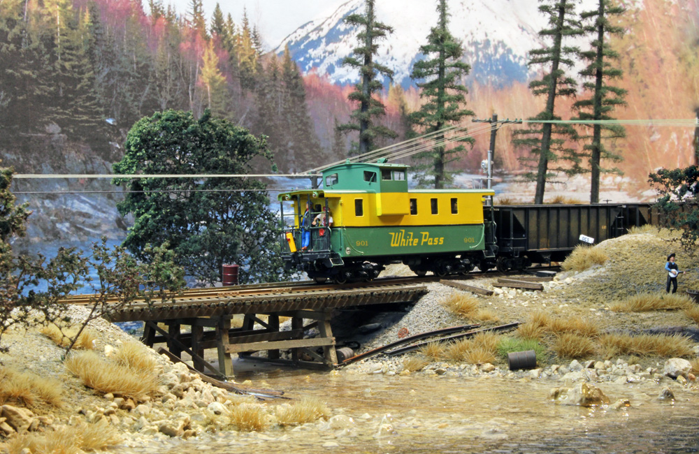 Railfans ride a green-and-yellow caboose behind a coal train as it crosses a short wooden trestle over a creek