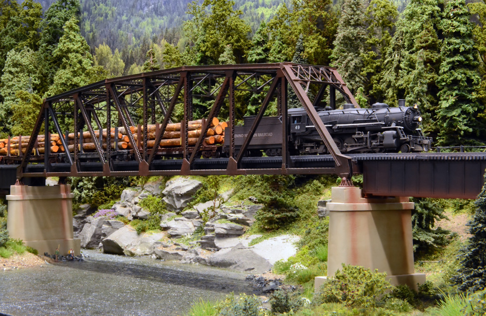 A steam locomotive pulls a string of loaded log cars over a steel trestle above a creek in a forest