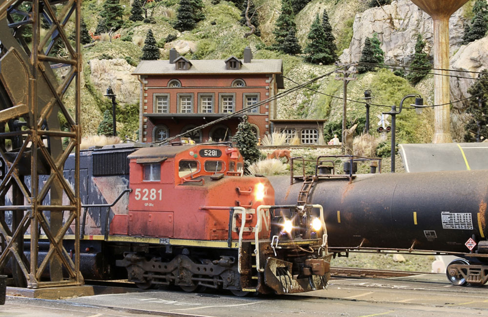 A weathered red-and-black diesel idles alongside an ash tower and a tank car in a yard