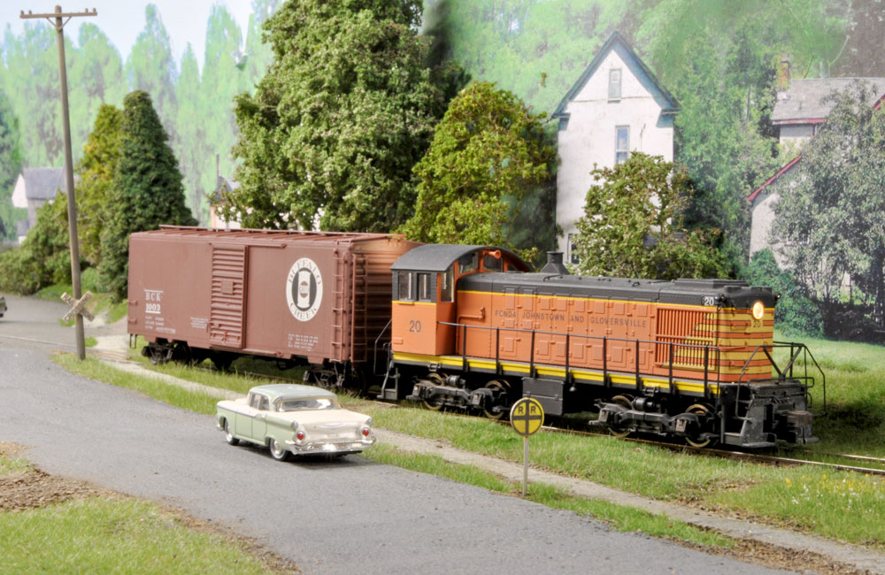 An orange-and-black diesel switcher pulls a boxcar alongside a rural road