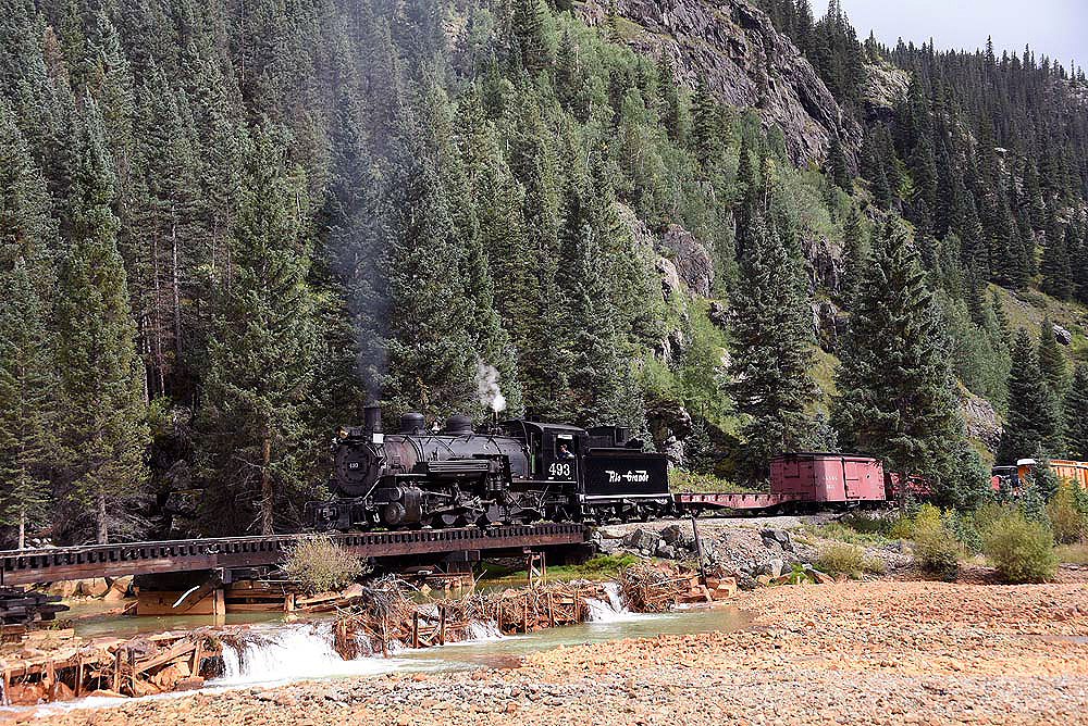 Narrow gauge steam train crossing a bridge in a forested scene.