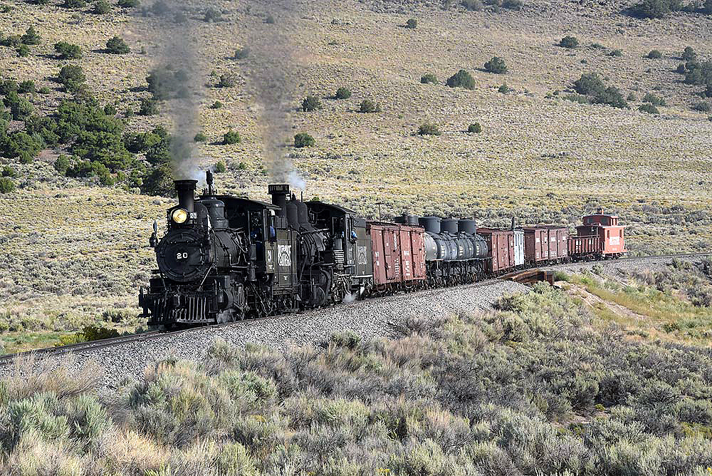 Two larger coal-fired steam locomotives lead a freight train through scrub and grassland.
