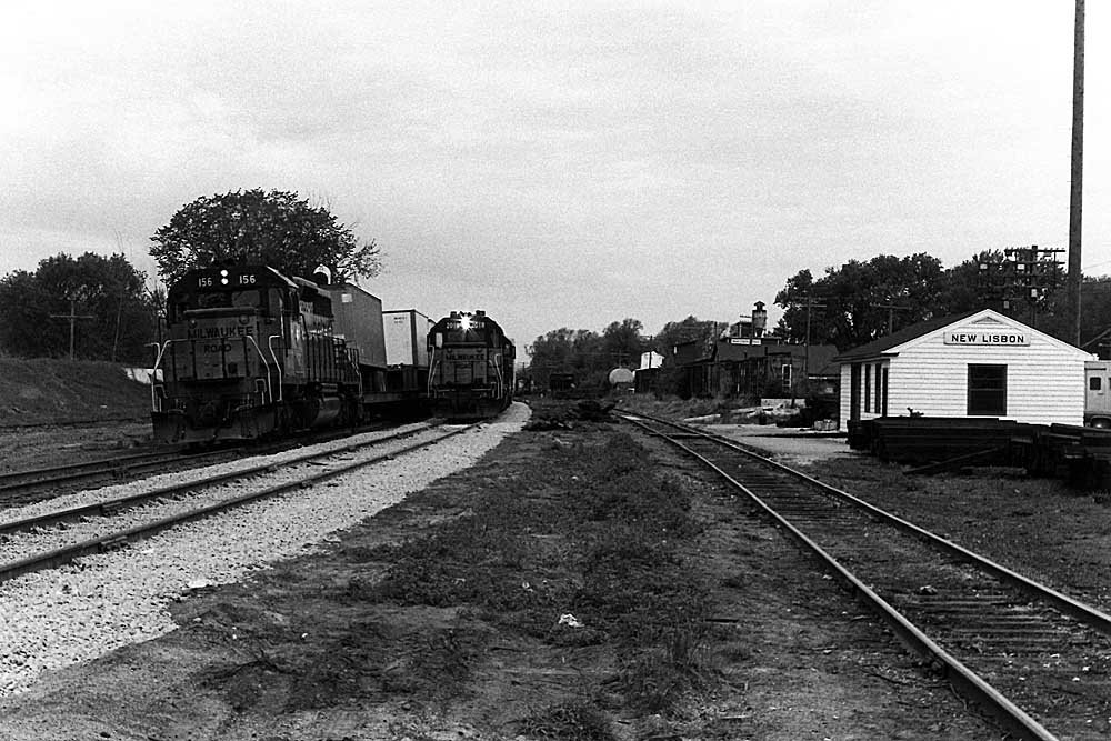 Two diesel-powered freight trains appear side-by-side on double track in front of station