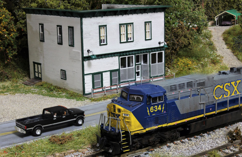 A gray, blue, and yellow modern diesel rolls past a two-story white clapboard country store