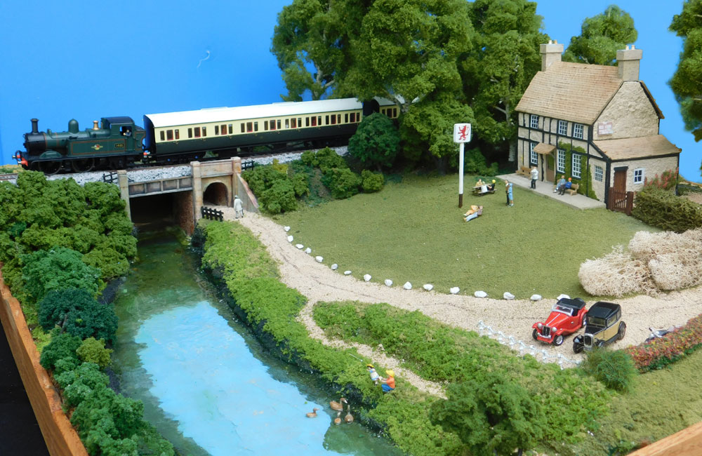A British tank locomotive pulls a pair of coaches past people relaxing in the garden of a country pub alongside a river