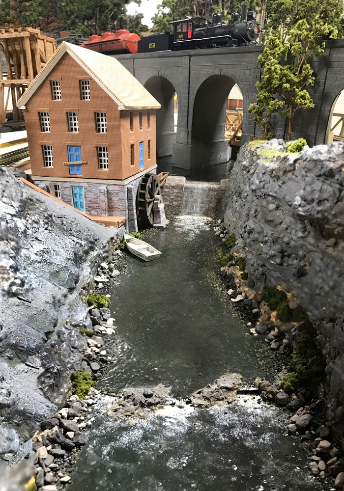 A steam engine pulls a train over a stone arch bridge above a river with a brick mill powered by a water wheel