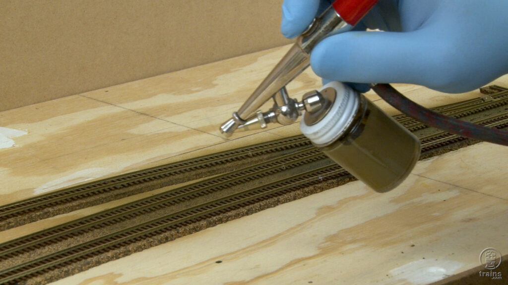 A gloved hand holding an airbrush filled with brown paint