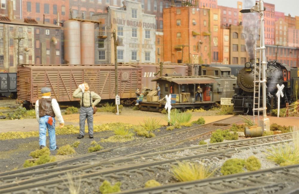 Two figures stand trackside while their steam locomotive rests by the yard office