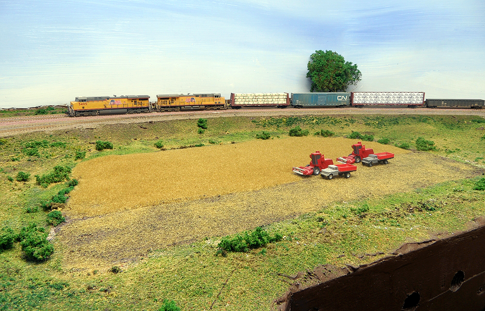 A yellow Union Pacific locomotive-led train passes a field with red farm equipment in N Scale.