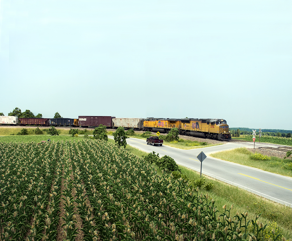 A yellow Union Pacific locomotive leads a freight train passed corn fields. On an HO scale layout.
