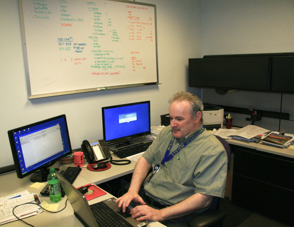 Man at desk in office with whiteboard and two computer screens. He managed Amtrak trains during the holidays.