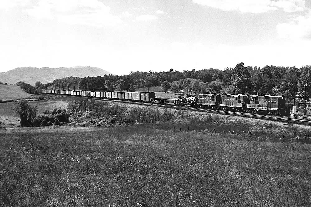 Three diesel locomotives move train of mostly boxcars through wooded hills
