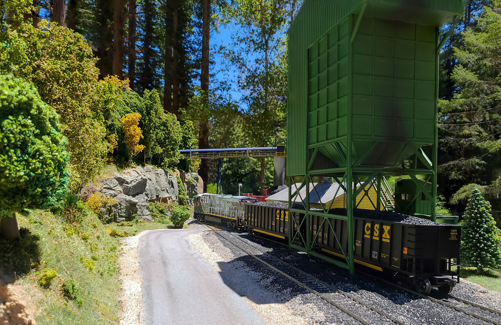 A modern diesel in a military camouflage paint scheme moves coal gondolas under a green coal loader