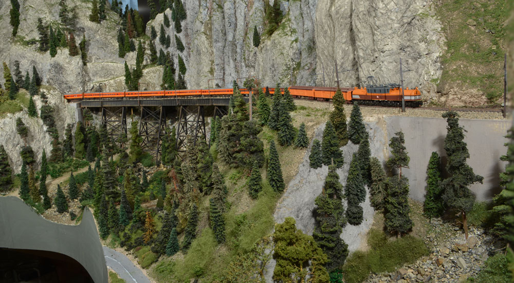 A maroon-and-orange passenger train behind an electric locomotive snakes along a rocky slope