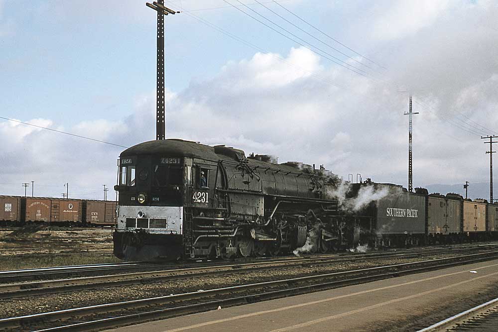 Steam locomotive with cab in front pulls freight train through yard