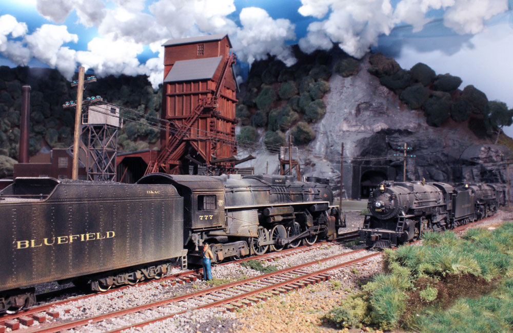 A trainman stands alongside a steam locomotive on a siding and watches another train approach on the main