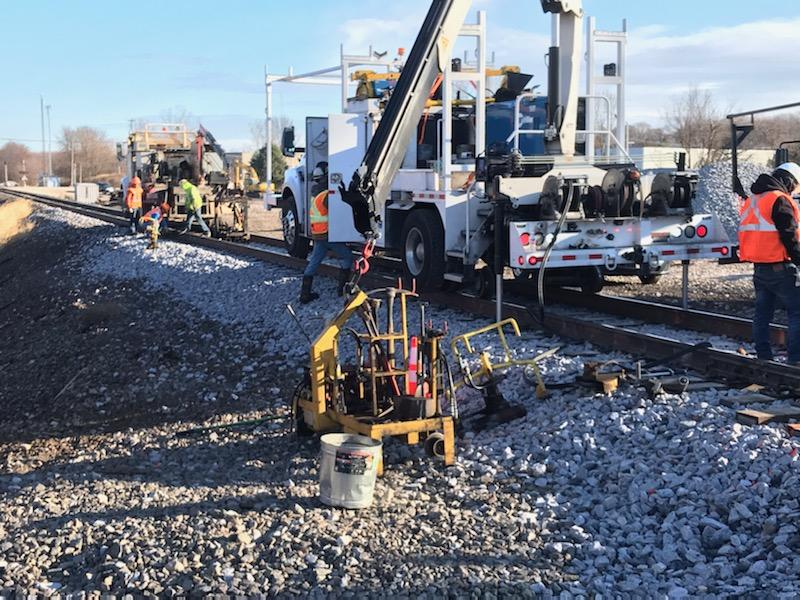 A white heavy-duty high-rail truck with a crane sits on the single-track Canadian National main line as orange-vest-clad workers stand along the rails and a yellow piece of welding equipment sits in the foreground.