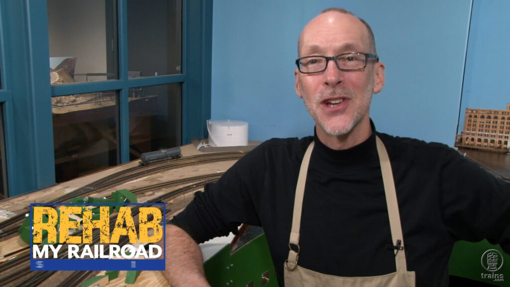Man standing near an unfinished model train layout in an episode that will discuss printed backdrop installation.
