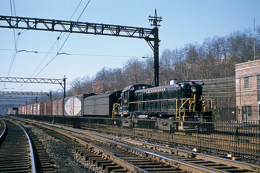 Black diesel locomotive pulls freight trains through junction under catenary wires