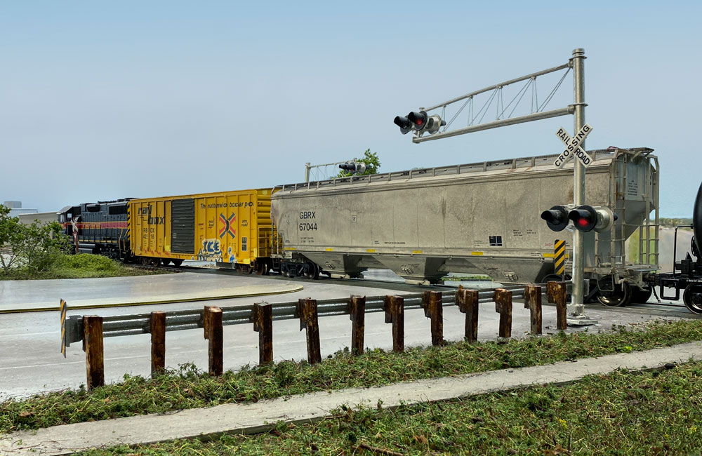 A black-and-red diesel pulls two freight cars across a grade crossing as warning lights flash.