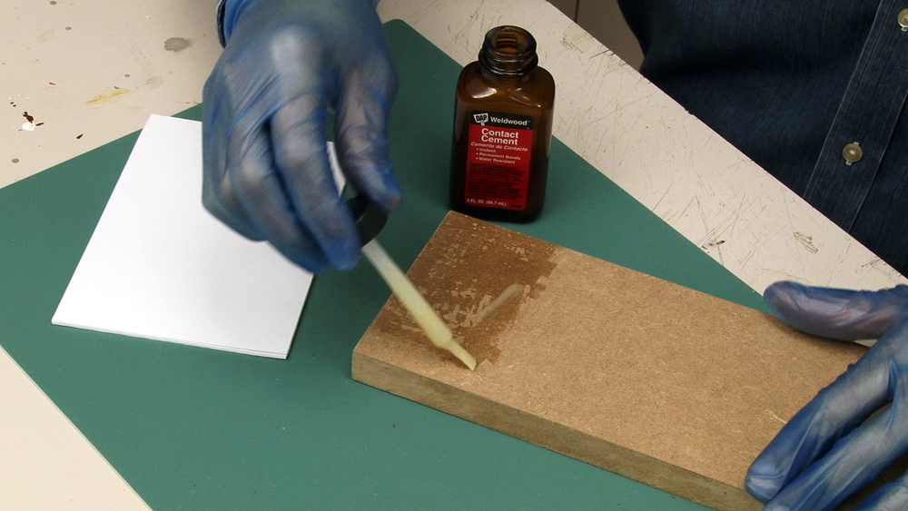 Rubber-gloved hands apply contact cement with a brush to dissimilar materials on a workbench surface