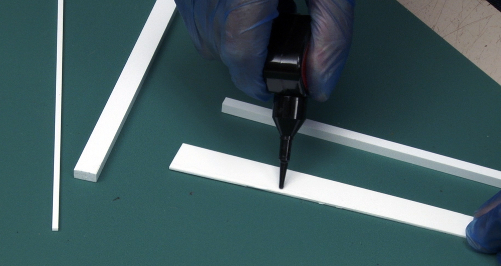 A rubber-gloved hand applies liquid cement to plastic strips on a workbench