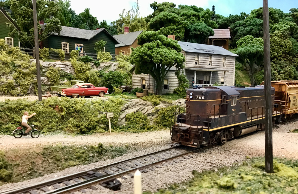 A boy on a bicycle riding down a dirt road waves to the engineer of a passing train