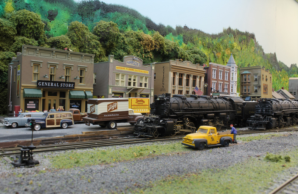 A large black steam locomotive waits while another pulls out of a yard in a mountain town
