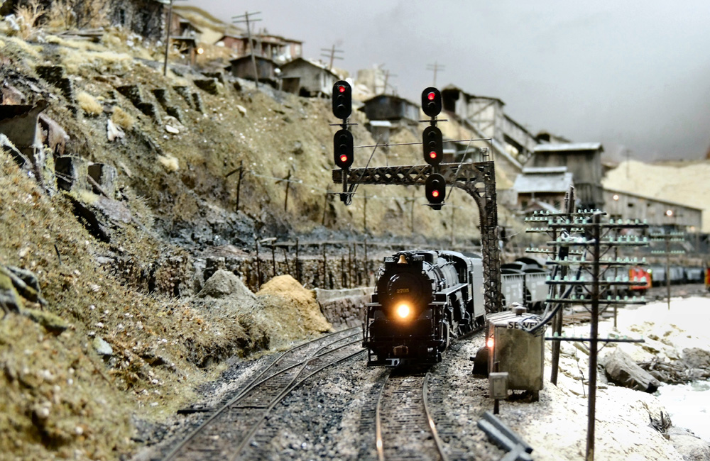 A steam locomotive with a coal train rolls under a signal bridge with a coal tipple in the distance