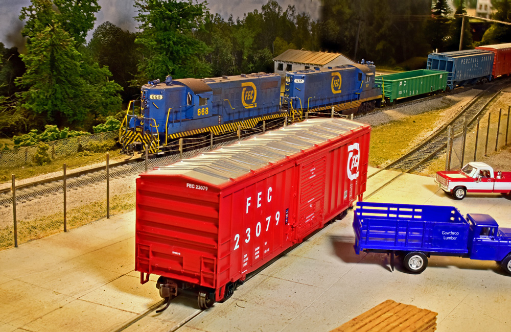 A bright red boxcar sits in a paved lot next to a blue stakebed truck while a train passes behind