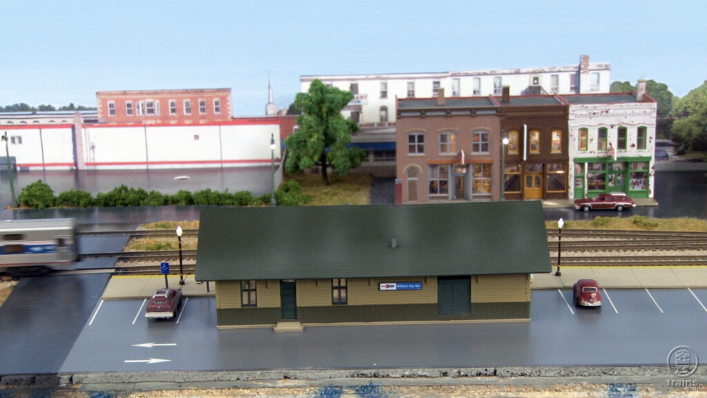 An N scale Amtrak depot with cars parked on each side and buildings in the background