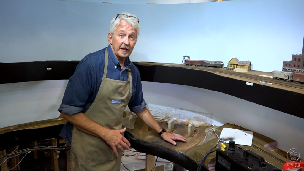 Man standing over a model train layout wearing an apron.
