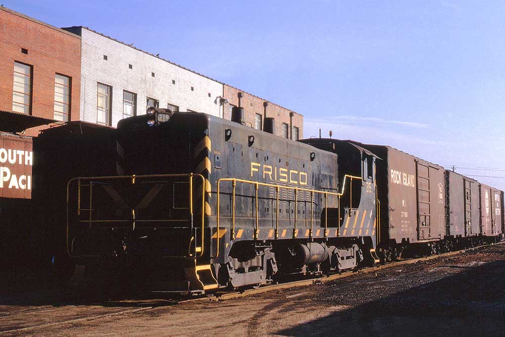 Black and gold diesel locomotives with Frisco freight train among industrial buildings