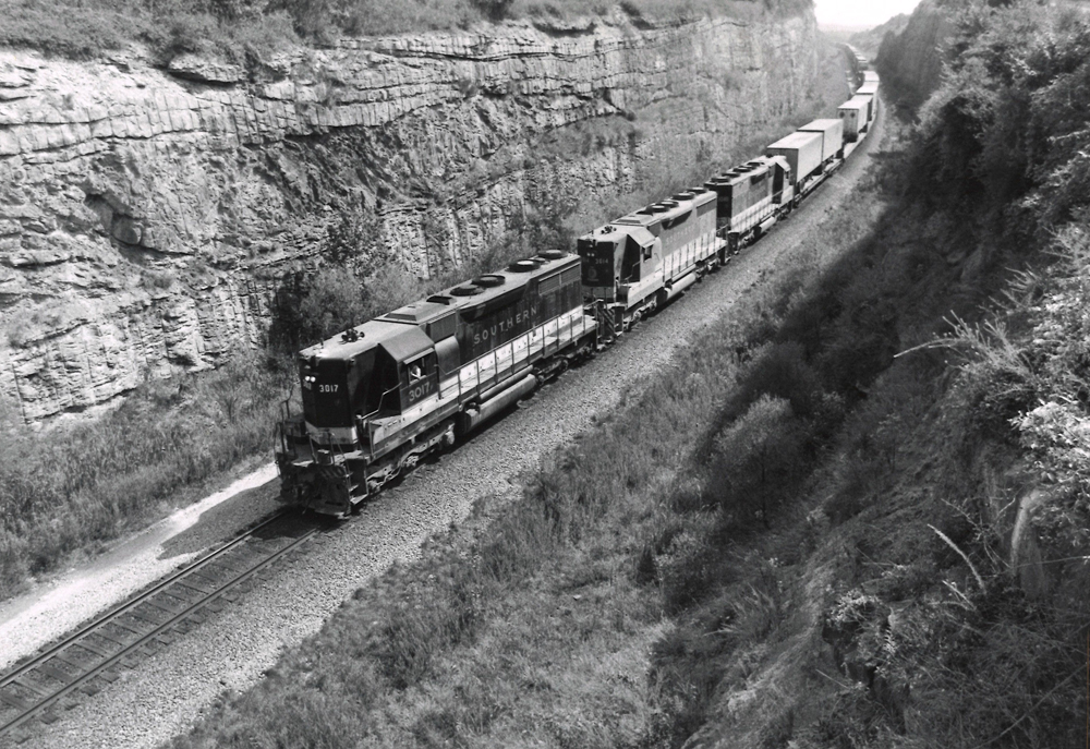 Black diesel locomotive leads a freigh train through a rock cut.