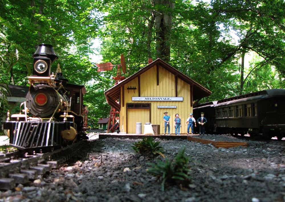 Garden railway scene with a steam locomotive, depot, and several figures