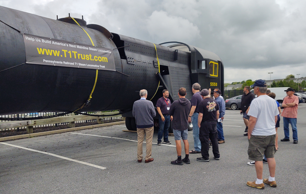 Man standing next to partially built locomotive talking to spectators