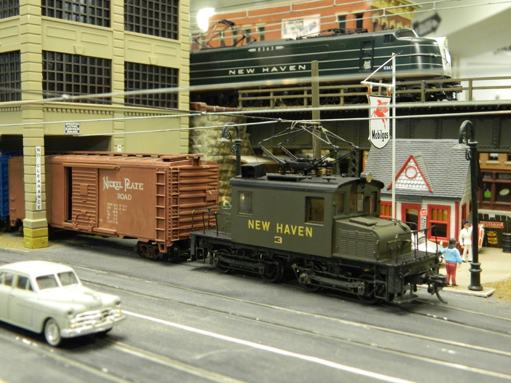 A green New Haven electric locomotive pushes a boxcar into a covered loading dock while in the background a larger one crosses a bridge.
