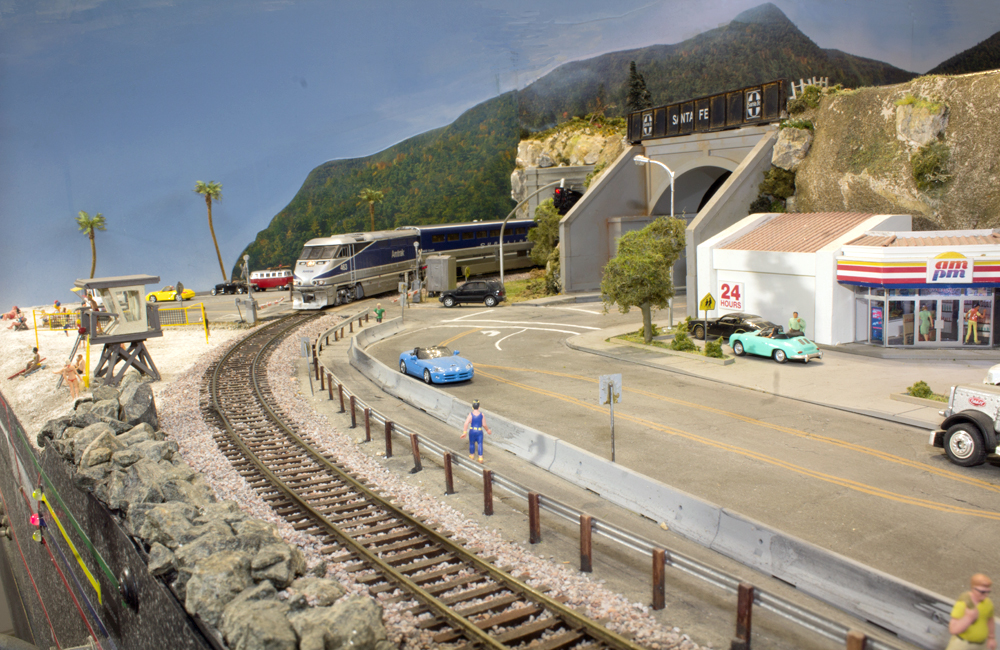 A blue-and-silver Amtrak train emerges from a tunnel and curves between a beach and a highway with a convenience store nearby