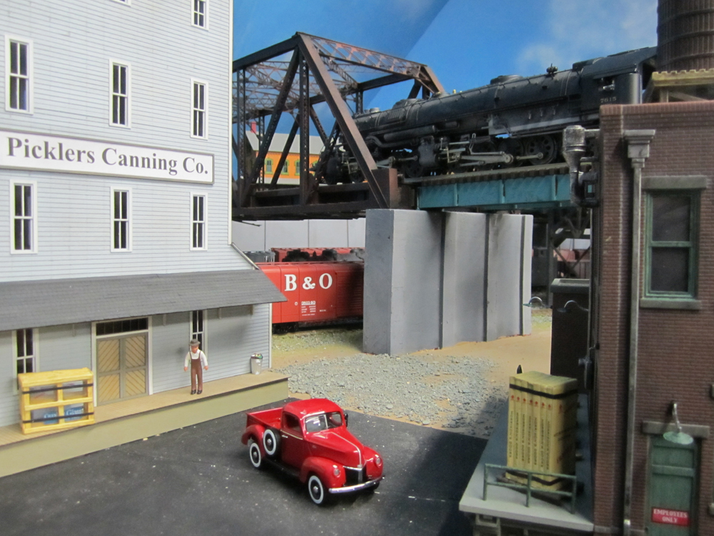 An articulated steam locomotive rumbles across a steel trestle above a street in an industrial area
